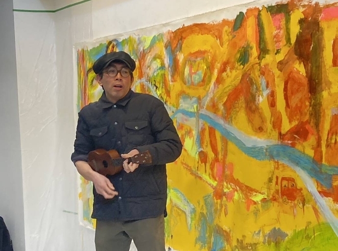 写真：辻ポポロン元彦さんが、絵の前で弾き語りしている